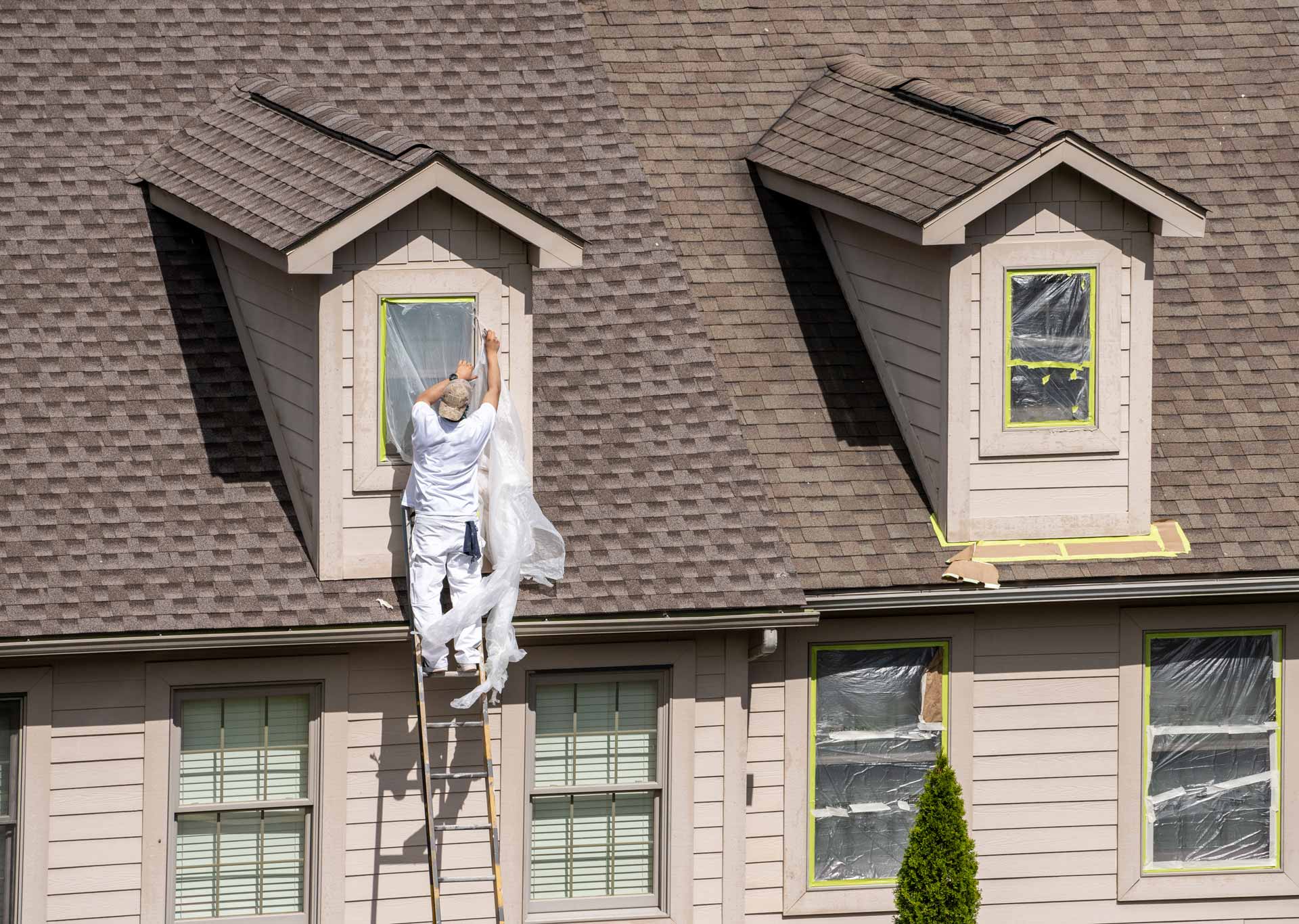 Painting contractor on ladder secures plastic coverings on building's windows